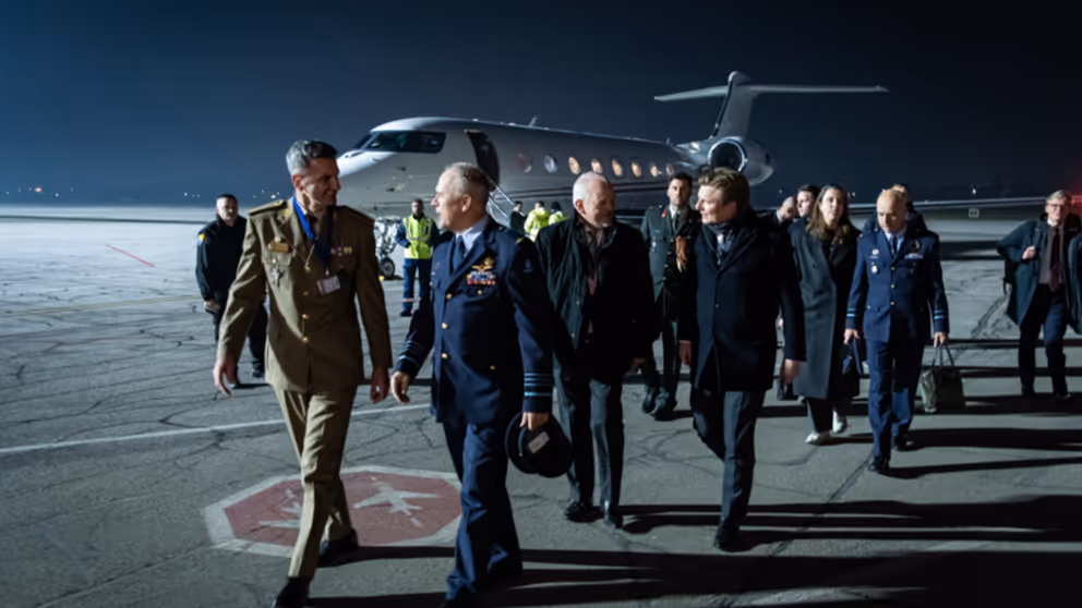 De CDS en minister Brekelmans lopen met een delegatie op een donkere landingsbaan. Op de achtergrond is een vliegtuig te zien.