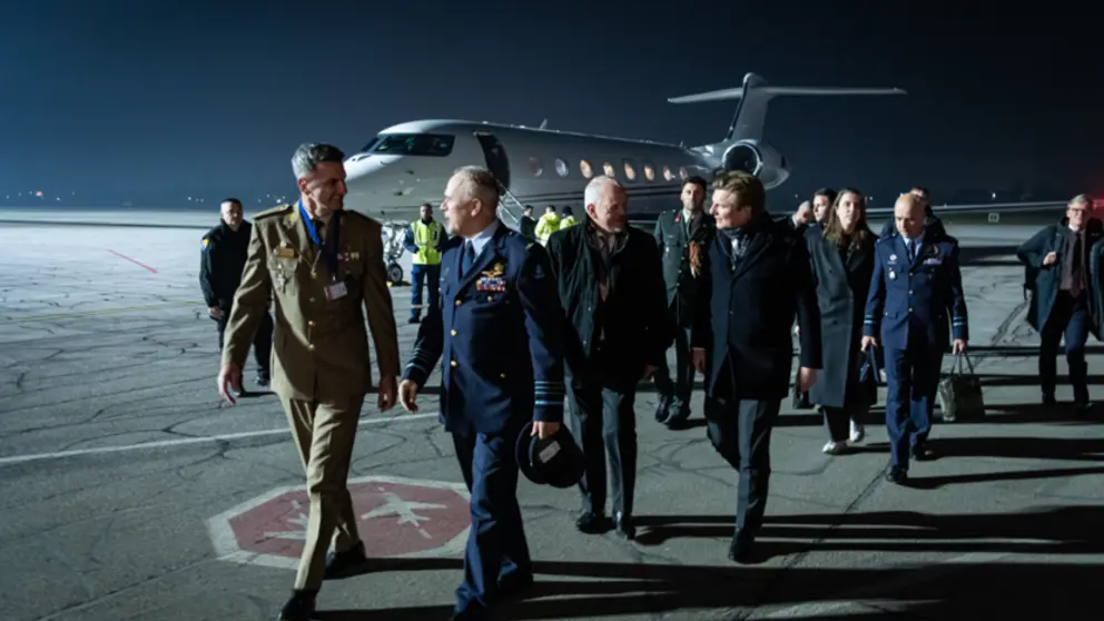 De CDS en minister Brekelmans lopen met een delegatie op een donkere landingsbaan. Op de achtergrond is een vliegtuig te zien.