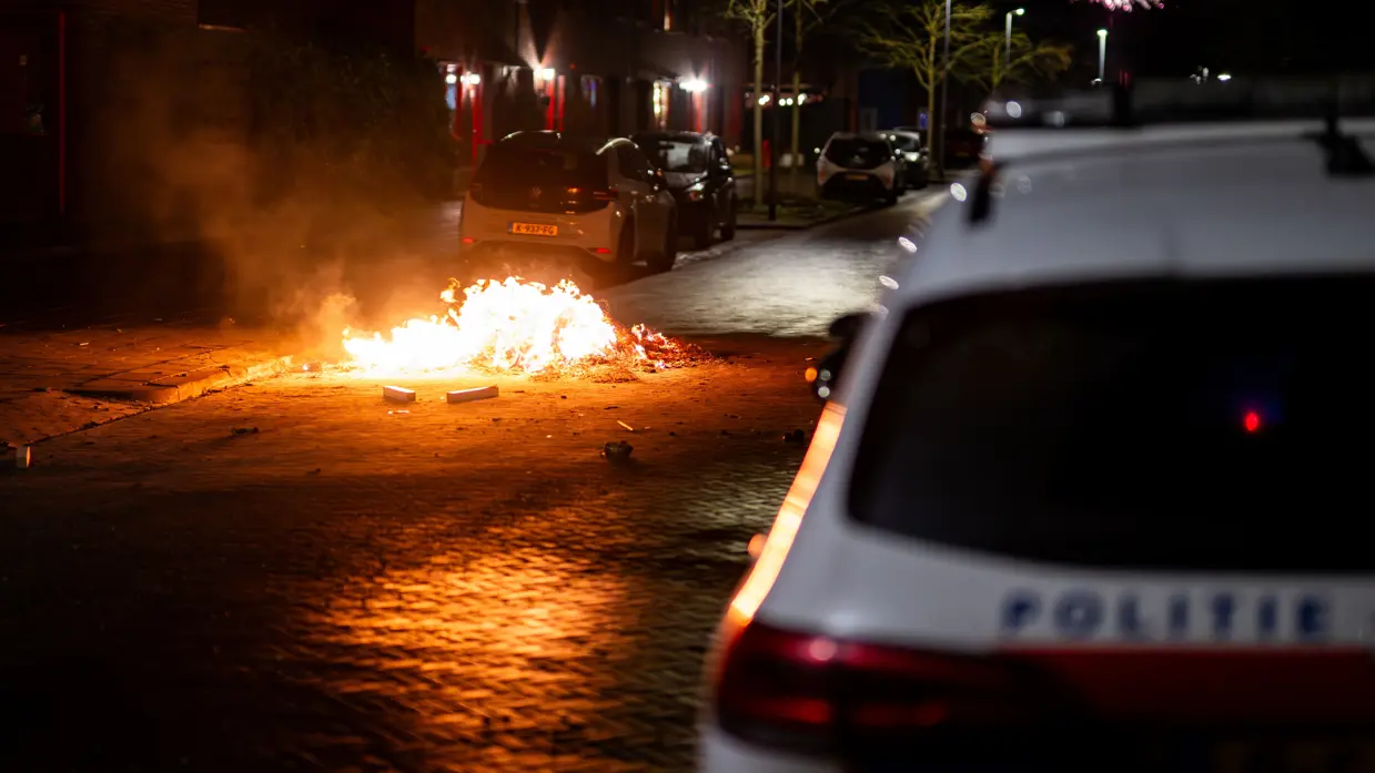 Voertuig van de politie met vlammend vuurwerk op de achtergrond.