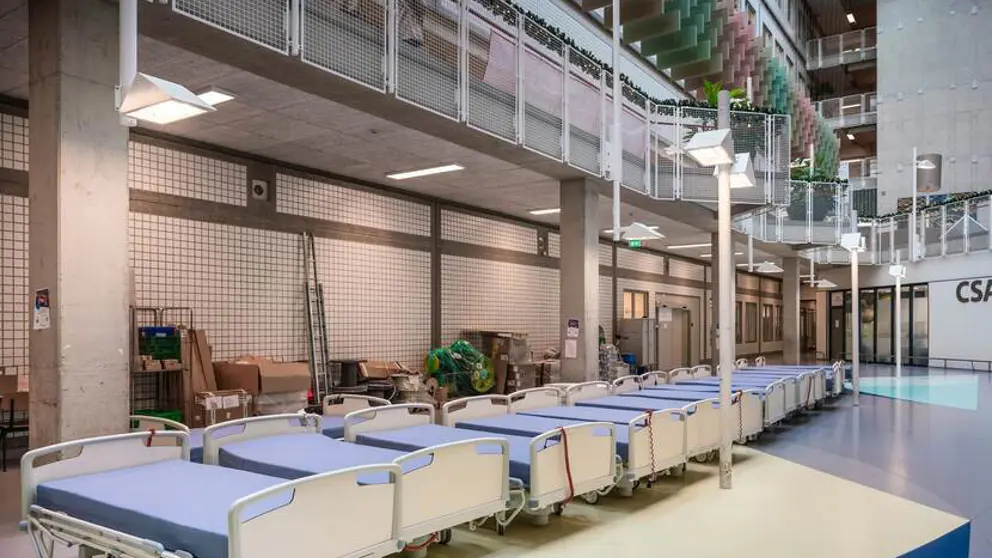 Een dertiental bedden met blauwe matrassen staan in de hal van een ziekenhuis.