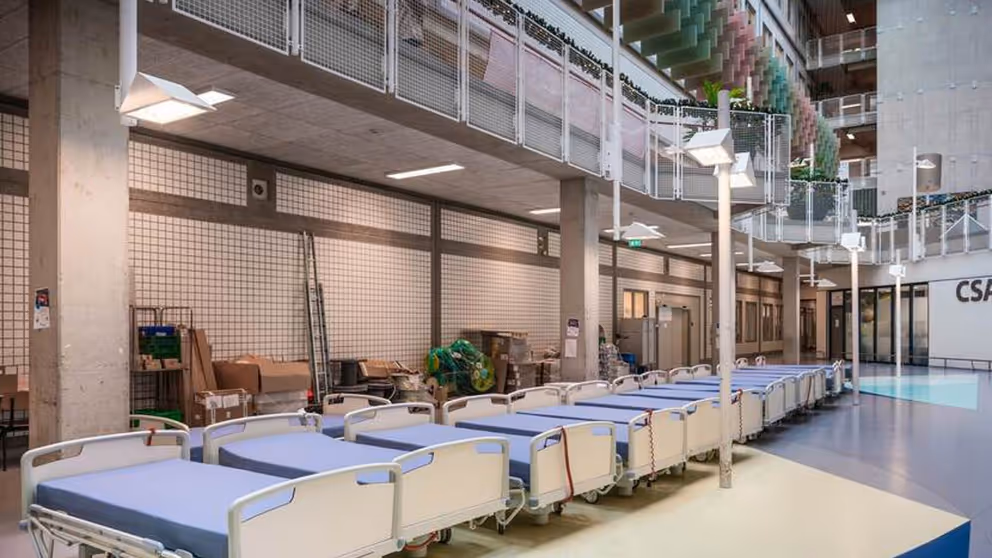 Een dertiental bedden met blauwe matrassen staan in de hal van een ziekenhuis.
