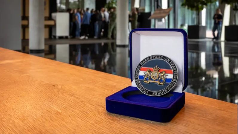 Coin staand in een opengeklapt doosje op een tafel. In het midden van de coin staat de Nederlandse vlag met daar overheen het Wapen van Nederland. Op de rand staat de tekst ‘Defensie dankt haar stage- en praktijkbegeleiders’.