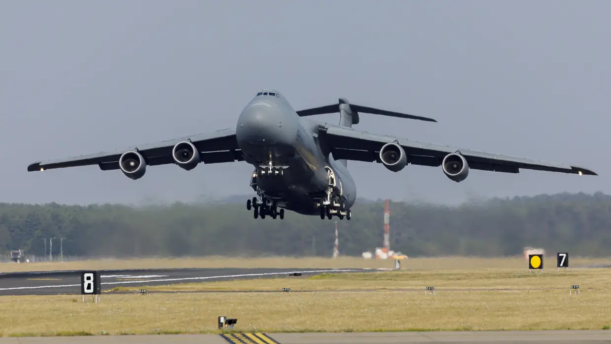 Groot grijs militair vliegtuig met vier motoren vliegt boven een startbaan, omringd door gras en bomen op de achtergrond.