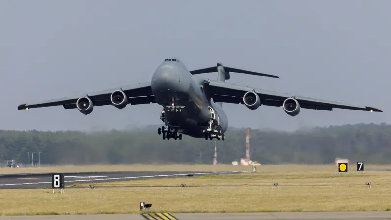 Groot grijs militair vliegtuig met vier motoren vliegt boven een startbaan, omringd door gras en bomen op de achtergrond.