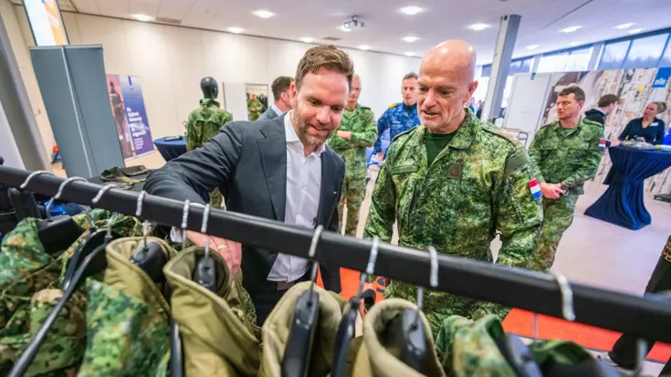 Staatssecretaris Derk Boswijk, in pak, gaat met zijn hand door een kledingrek met militaire uniformen. Een militair kijkt met hem mee.