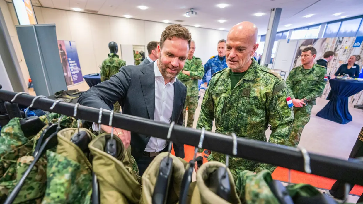 Staatssecretaris Derk Boswijk, in pak, gaat met zijn hand door een kledingrek met militaire uniformen. Een militair kijkt met hem mee.
