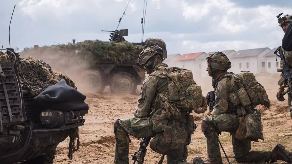 2 militairen in volledige gevechtsuitrusting beiden op een knie tussen rook. Voor hen 2 militaire voertuigen.