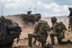2 militairen in volledige gevechtsuitrusting beiden op een knie tussen rook. Voor hen 2 militaire voertuigen.