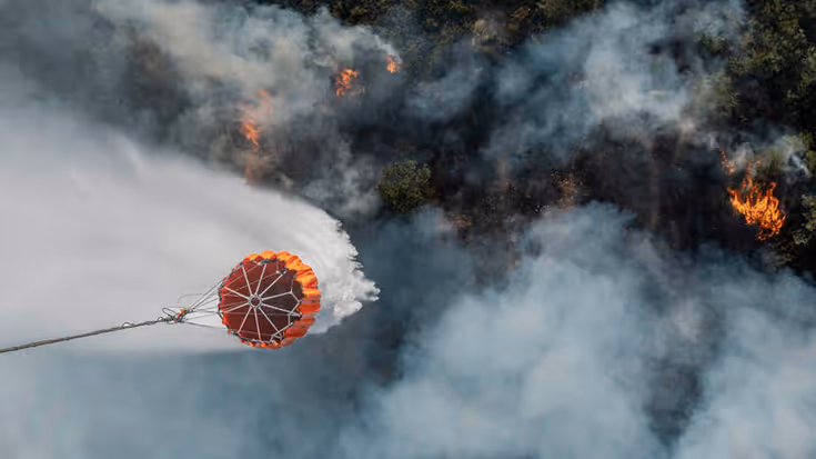 Foto vanuit Chinook-helikopter recht naar beneden. Een bambi bucket heeft zojuist water gedropt op brandend terrein.