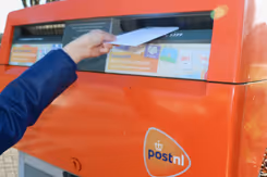 Hand gooit witte enveloppe in rode brievenbus.