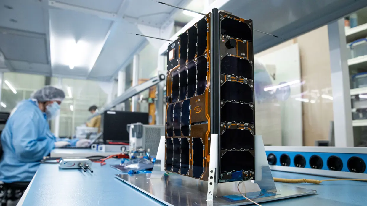 Foto van de BRIK II-satelliet in productie met op de achtergrond een werknemer aan het werk.