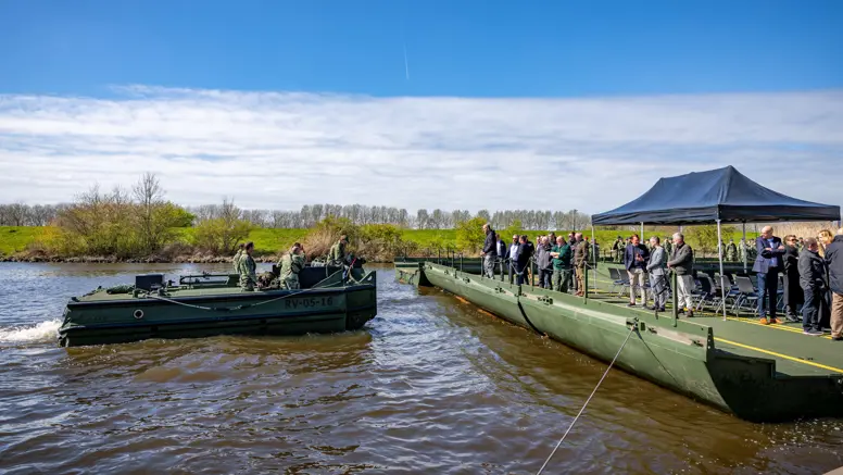 Een groepje militairen op een duwboot vaart richting een legergroen ponton. Daarop staan toeschouwers. Daarboven een zonnige lucht.