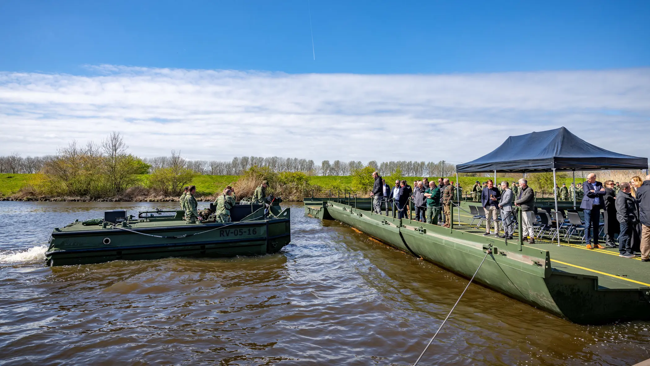 Een groepje militairen op een duwboot vaart richting een legergroen ponton. Daarop staan toeschouwers. Daarboven een zonnige lucht.