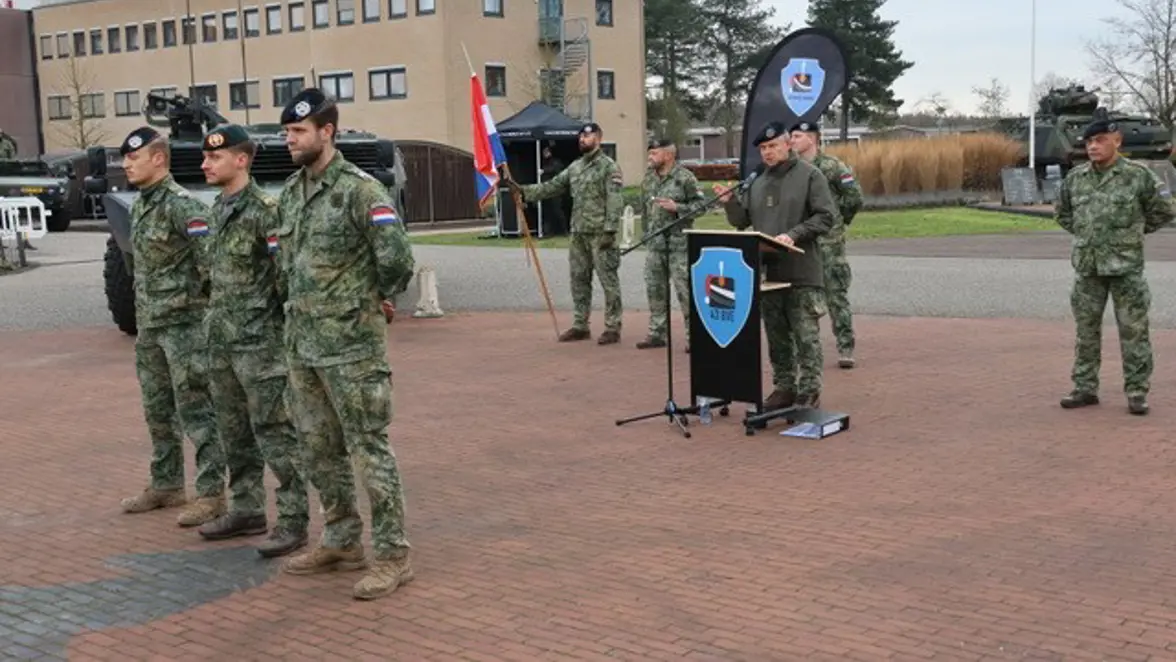 Militairen in uniform tijdens een buitenplechtigheid. Een persoon spreekt achter een lessenaar, met een Nederlands en een eenheidsvaandel op de achtergrond.
