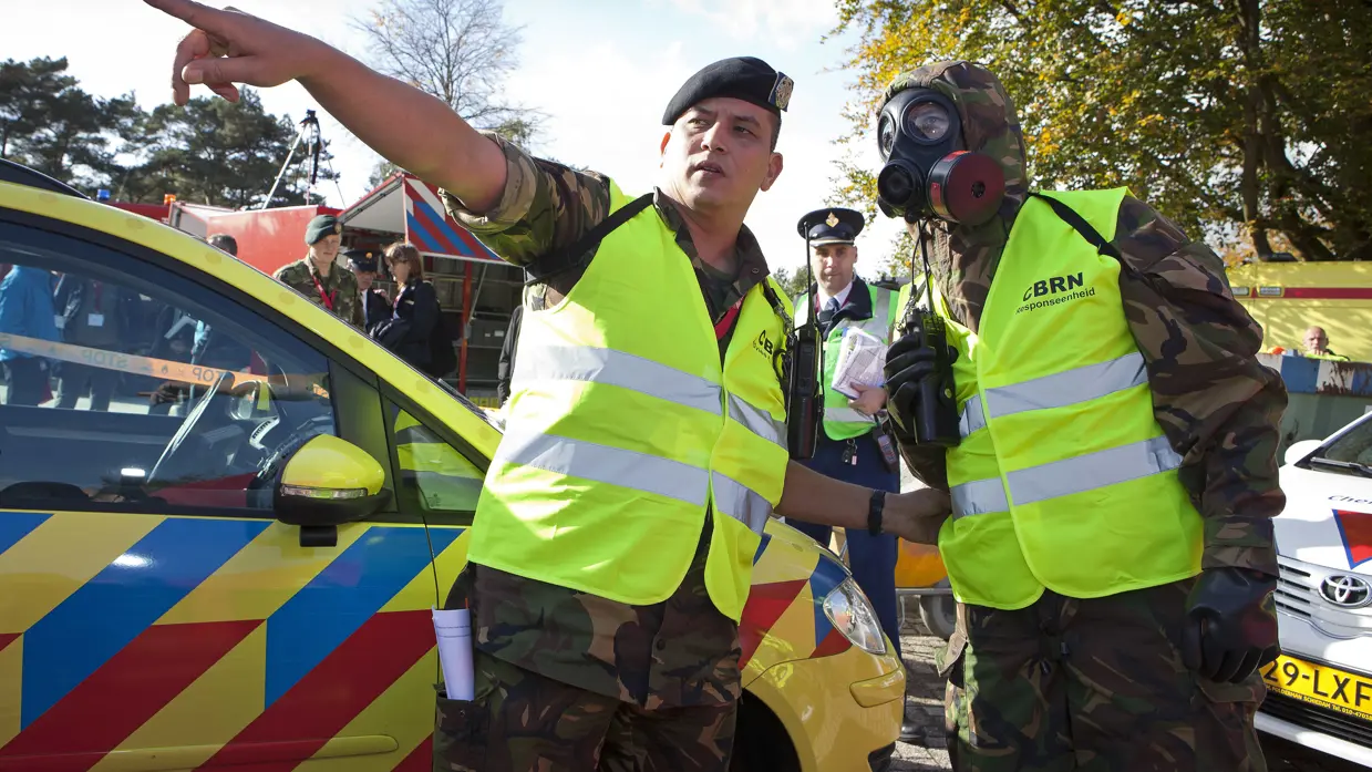 Een militair van de CBRN Respons Eenheid met beschermende uitrusting wordt naar een oefenramplocatie gewezen.