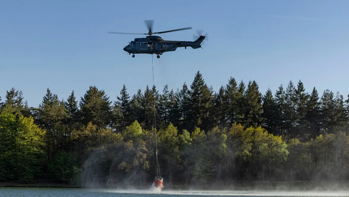 Een helikopter vliegt laag boven een meer en vult een oranje waterzak die onder de heli hangt. 