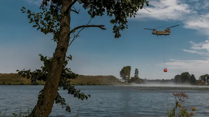 Een Chinook met Bambi Bucket vliegt boven de Maas. Op de voorgrond een boom.