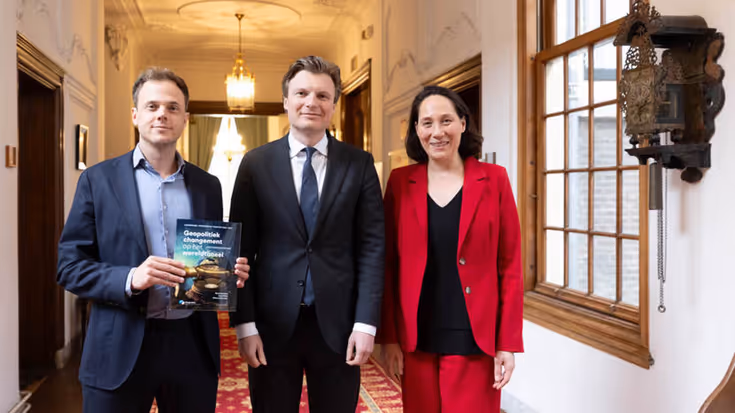 Twee mannen in pak en een vrouw in het rood staan naast elkaar. De man links houdt een rapport vast.