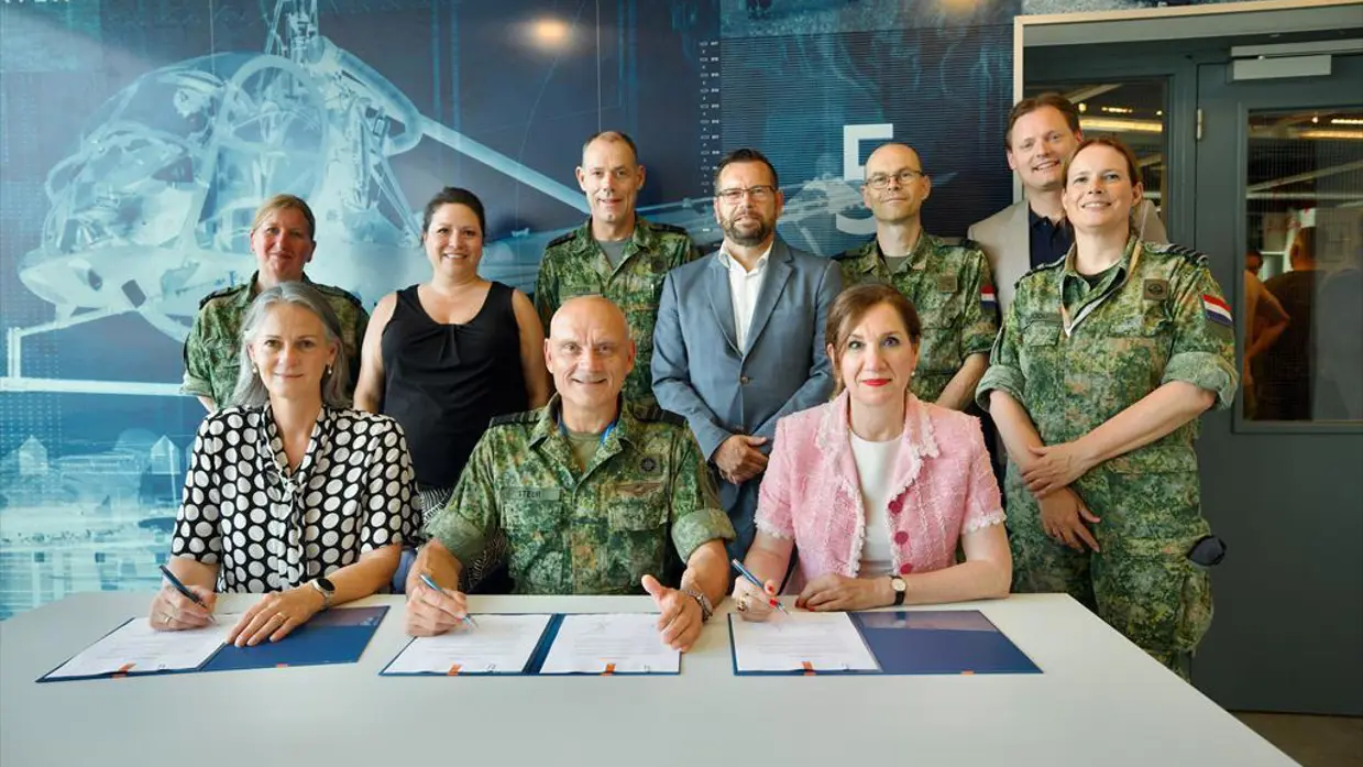 Generaal Steur zittend te midden van twee dames in burgerkleding. Alle drie hebben een pen in de hand en ondertekeningspapieren voor zich op tafel. Achter hen staan vier militairen in gevechtstenue en drie mensen in burgerkleding.