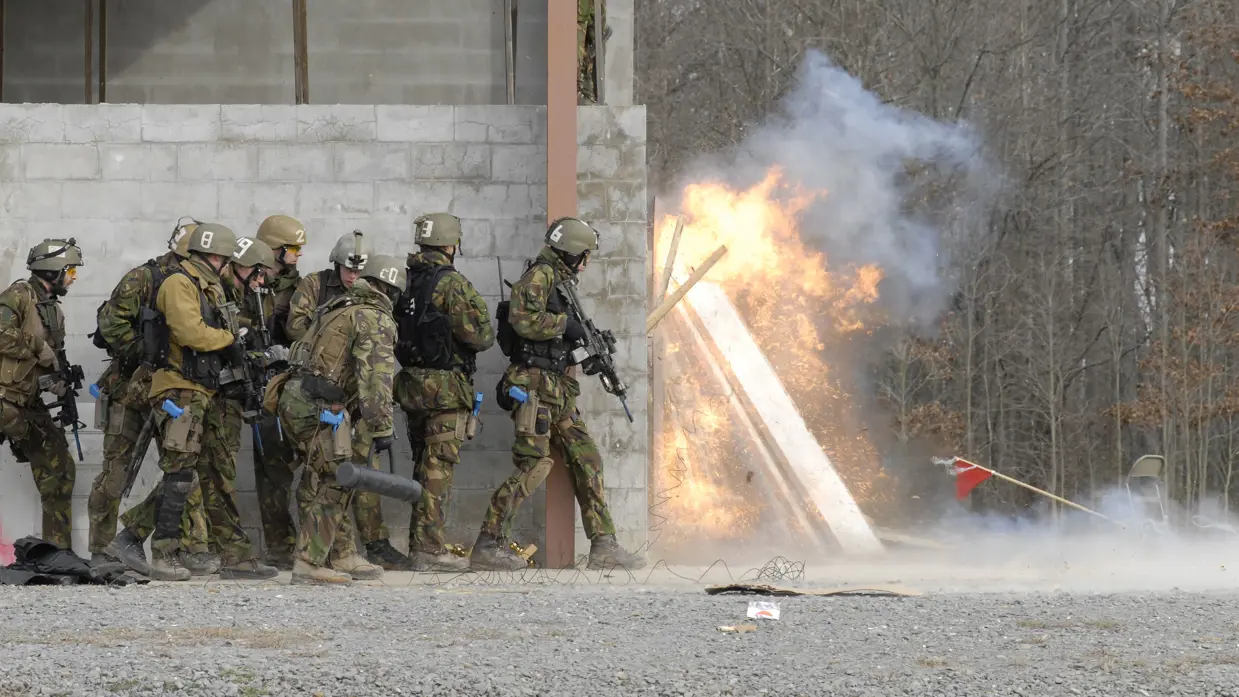 Militairen staan naast muur van oefengebouw terwijl de deur explodeert om de hoek.
