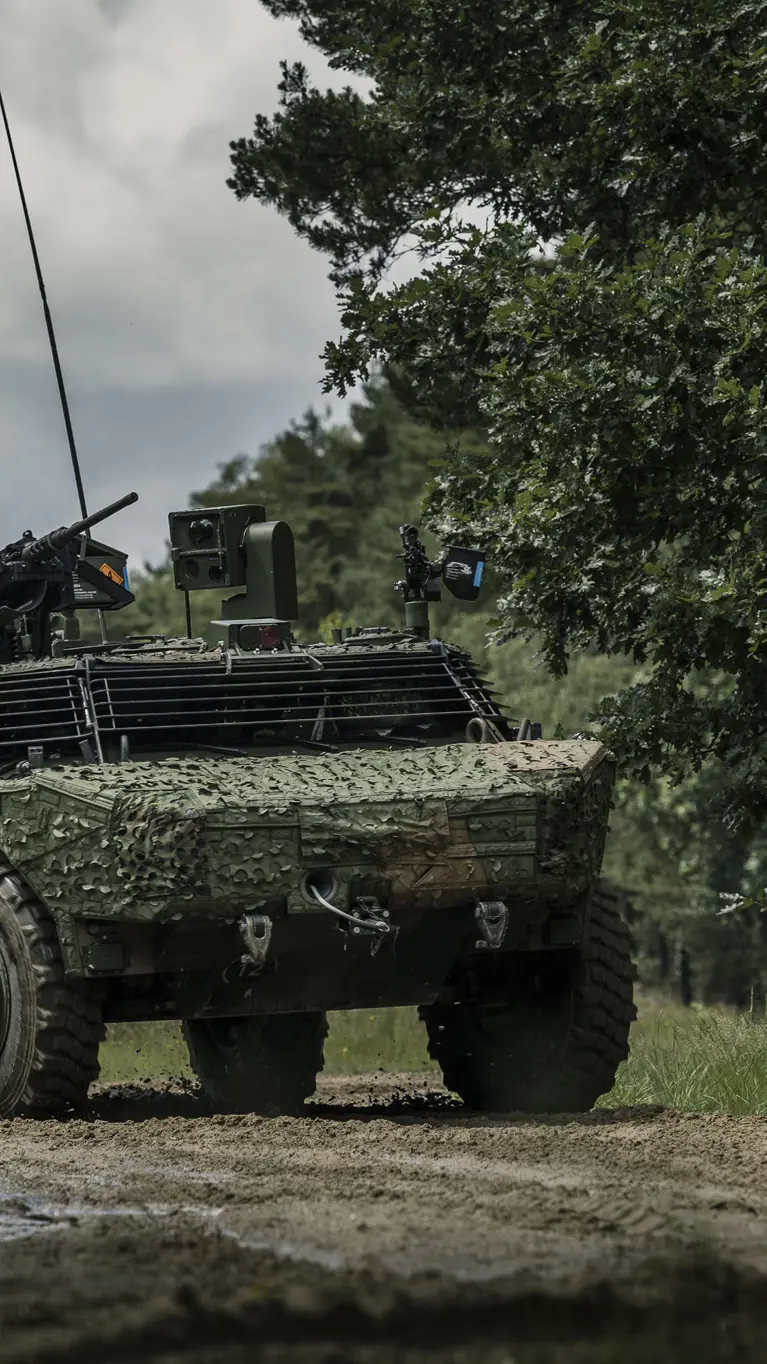 Een Fennek-voertuig met een camouflagenet rijdt door een beboste omgeving.