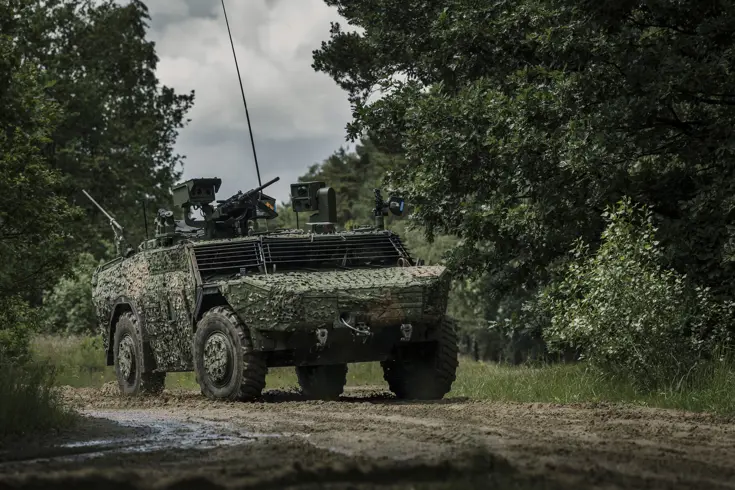 Een Fennek-voertuig met een camouflagenet rijdt door een beboste omgeving.