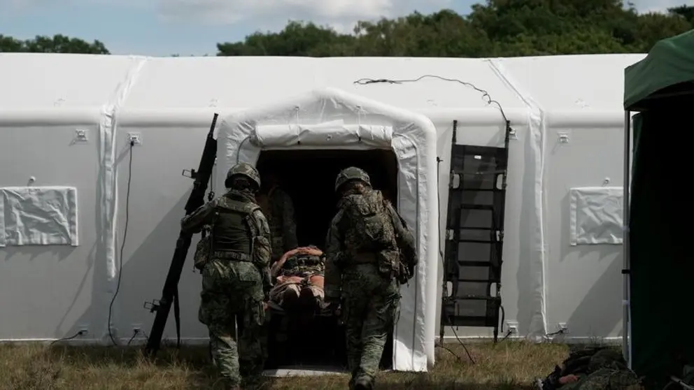 2 militairen op de rug gezien dragen een brancard met een gewonde een witte tent in.