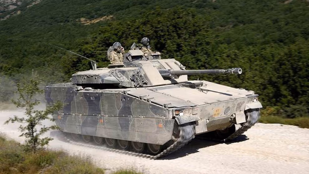 Een CV90 gevechtvoertuig rijdt op een zandpad. Op de achtergrond een groen begroeide heuvelrug.