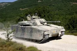 Een CV90 gevechtvoertuig rijdt op een zandpad. Op de achtergrond een groen begroeide heuvelrug.
