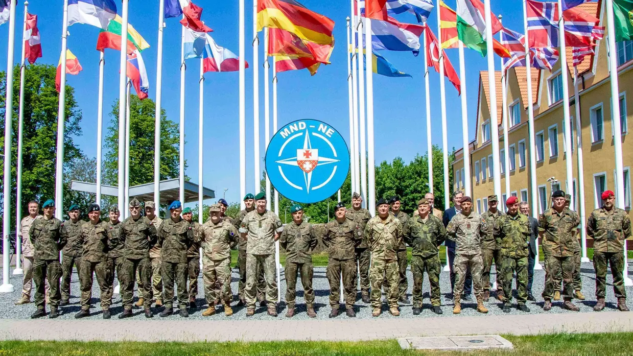 Een groep militairen staat, met de handen op de rug, opgesteld. Boven hen wapperen vlaggen van verschillende landen.