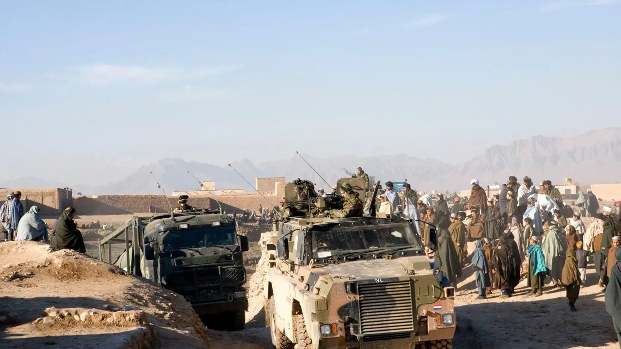 Archief 2007: Militaire voertuigen rijden over zandweg in Tarin Kowt. Mensen kijken toe.