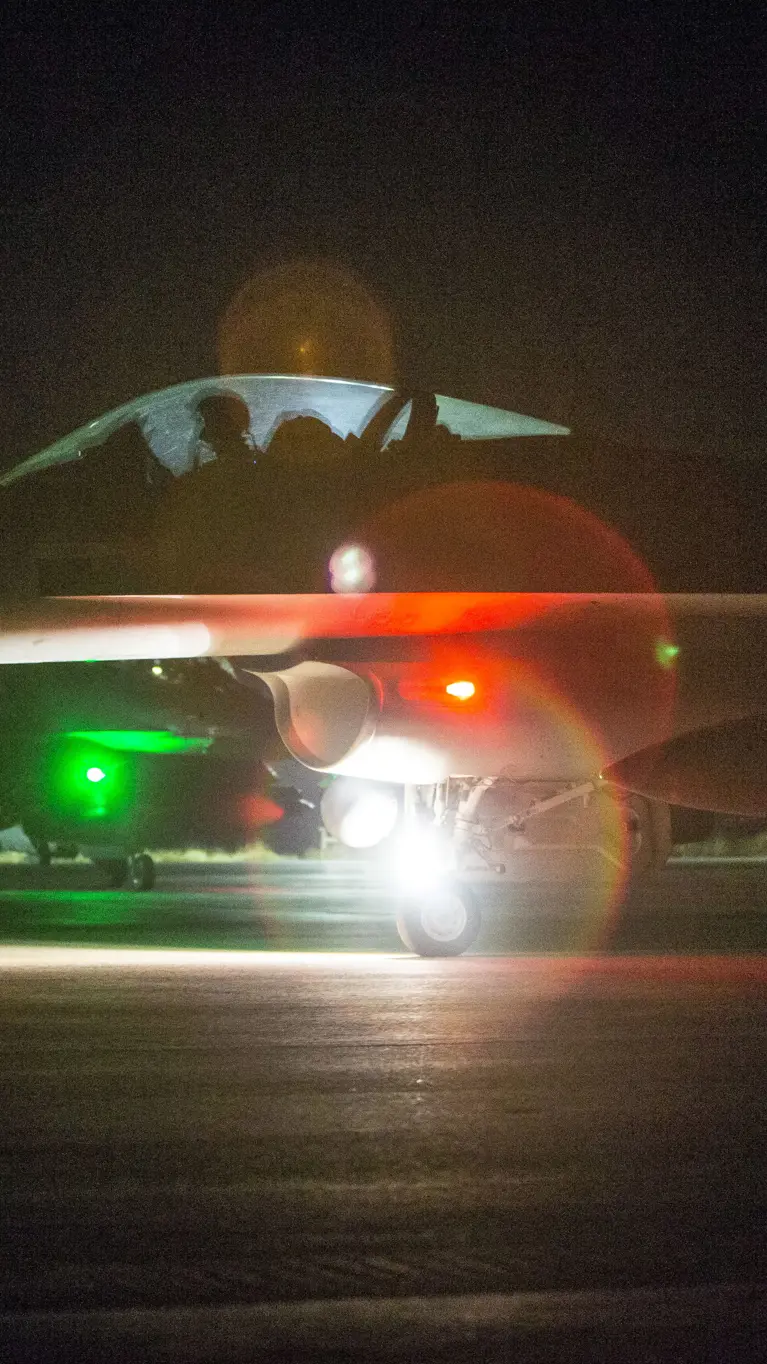 Nachtfoto van een taxiënde F-16 waarbij een groene en rode lamp fel in de camera schijnt.