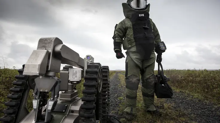 Een explosievenruimer van de EOD in het zware bommenpak en de explosievenrobot Telemax staan op het punt een gebouw binnen te gaan om een geïmproviseerd explosief te ruimen tijdens een oefening in IJsland (2016).