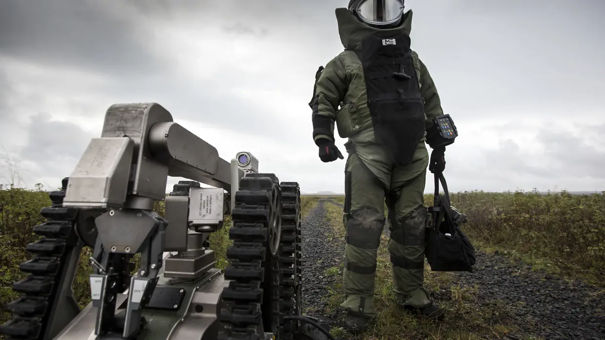Een explosievenruimer van de EOD in het zware bommenpak en de explosievenrobot Telemax staan op het punt een gebouw binnen te gaan om een geïmproviseerd explosief te ruimen tijdens een oefening in IJsland (2016).