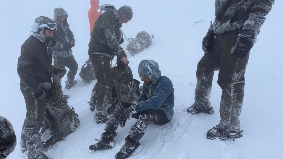 Mariniers staan in de sneeuw om een man heen die op de grond zit en uit een beker drinkt. 