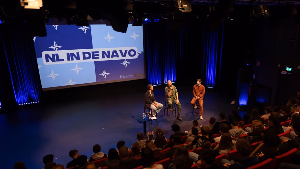 Mensen in een zaal en 3 personen op een stoel op een podium. Op de achtergrond groot scherm met de tekst: NL in de NAVO.