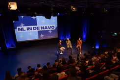 Mensen in een zaal en 3 personen op een stoel op een podium. Op de achtergrond groot scherm met de tekst: NL in de NAVO.