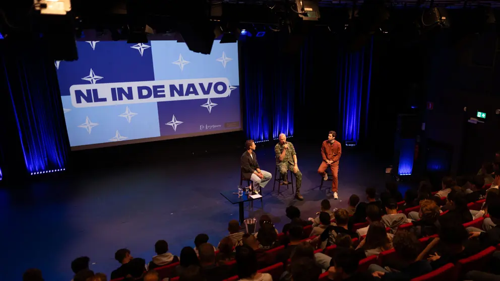 Mensen in een zaal en 3 personen op een stoel op een podium. Op de achtergrond groot scherm met de tekst: NL in de NAVO.