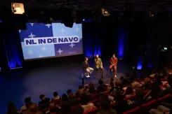 Mensen in een zaal en 3 personen op een stoel op een podium. Op de achtergrond groot scherm met de tekst: NL in de NAVO.
