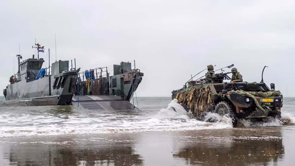LCU-landingsvaartuig ligt vlakbij het strand, en een Fennek-verkenningsvoertuig met 2 militairen erin rijdt op de voorgrond. 