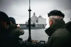 Doorkijk naar Zr.Ms. Johan de Witt op het water. Op de voorgrond een paal en een militair.
