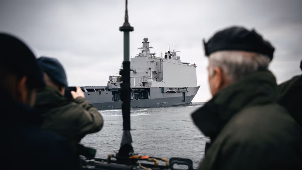 Doorkijk naar Zr.Ms. Johan de Witt op het water. Op de voorgrond een paal en een militair.