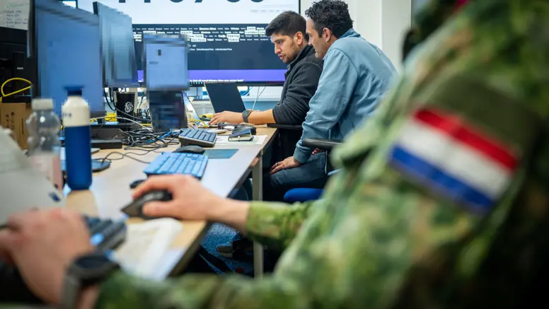 Drie mannen, van wie een Nederlandse militair, zitten achter hun PC aan een bureau met op de achtergrond een groot computerscherm. 