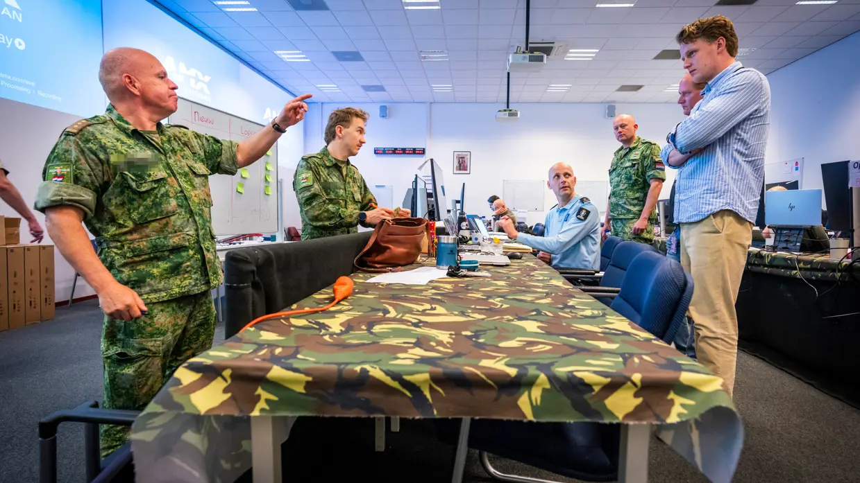 Een tafel met daaromheen zowel militairen als burgers. Kolonel Patrick wijst daarbij.