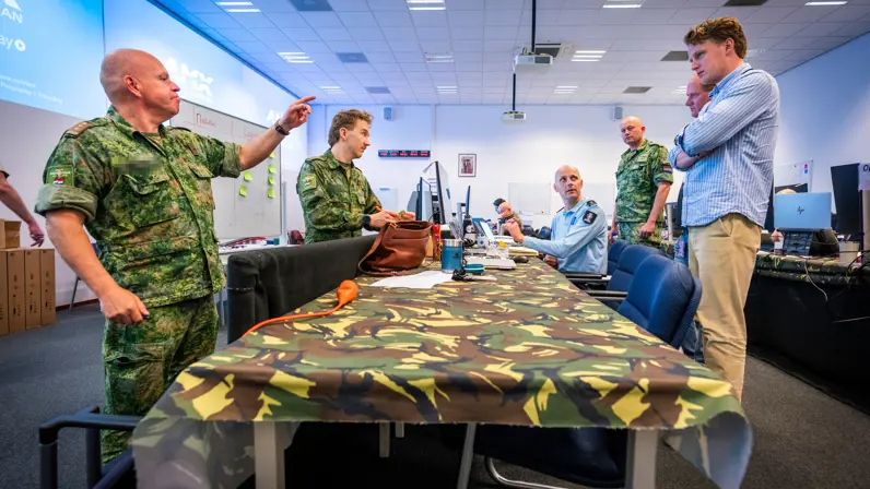 Een tafel met daaromheen zowel militairen als burgers. Kolonel Patrick wijst daarbij.