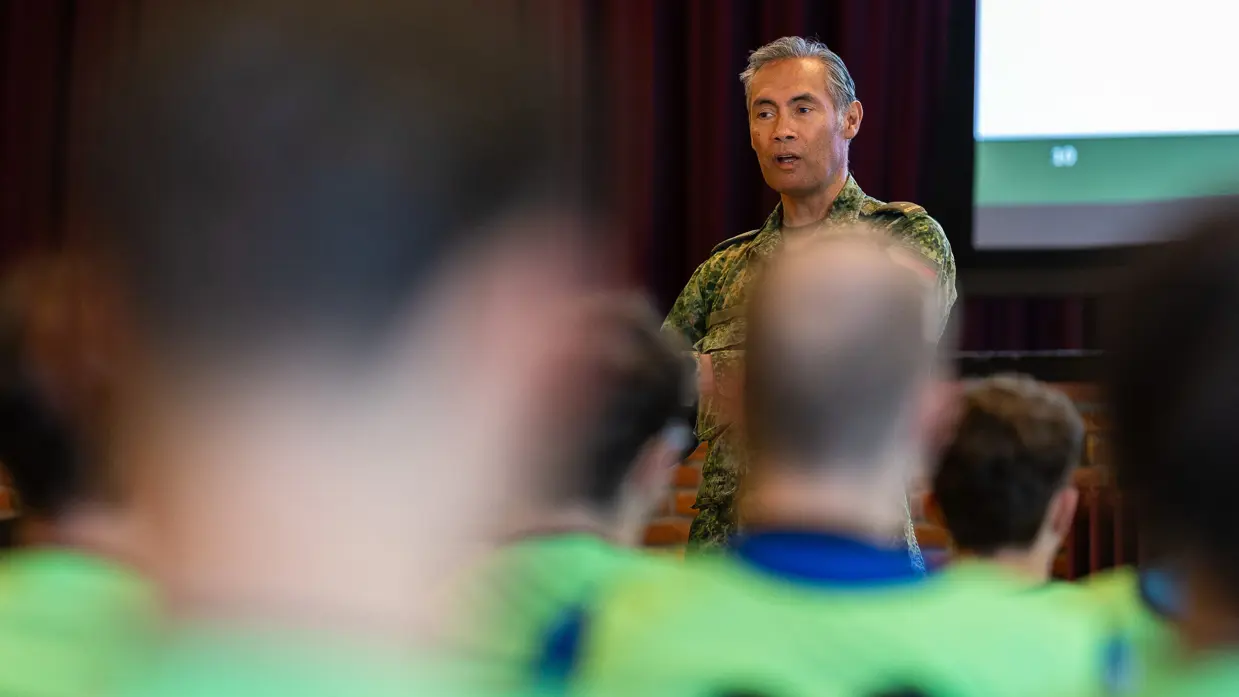 Een groep jongeren in gele vesten luistert in een zaal naar een militair die een presentatie geeft over organisatie en verschillende logo’s op een groot scherm.