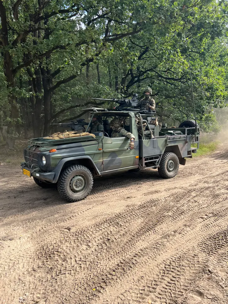 Een militaire jeep rijdt door een bosrijk zandpad, met soldaten erin en groene bomen als achtergrond. 