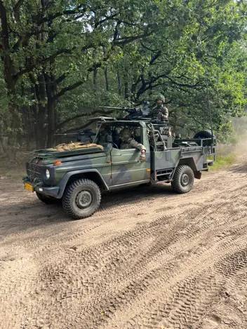 Een militaire jeep rijdt door een bosrijk zandpad, met soldaten erin en groene bomen als achtergrond. 