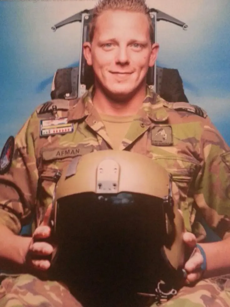 Sergeant Glenn Afman in vlekkenpak op een schietstoel met een vliegerhelm op zijn schoot.