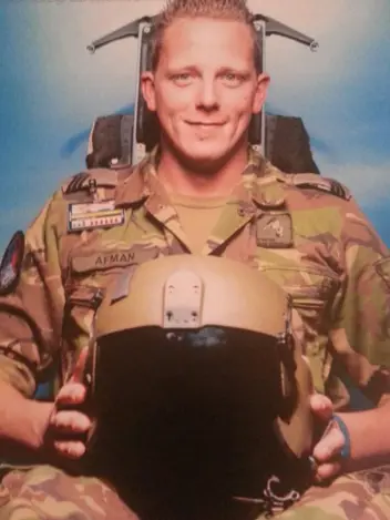 Sergeant Glenn Afman in vlekkenpak op een schietstoel met een vliegerhelm op zijn schoot.